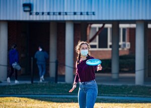 a woman student in a mask is playing frisbee outside her residence hall